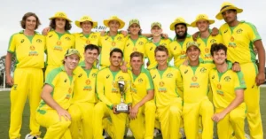 The image shows a group of Australian cricket players posing together outdoors in their bright yellow team uniforms with green accents. They are smiling and holding a trophy, celebrating their victory. The team is standing on a grass field with a clear sky in the background, showcasing a moment of camaraderie and achievement.